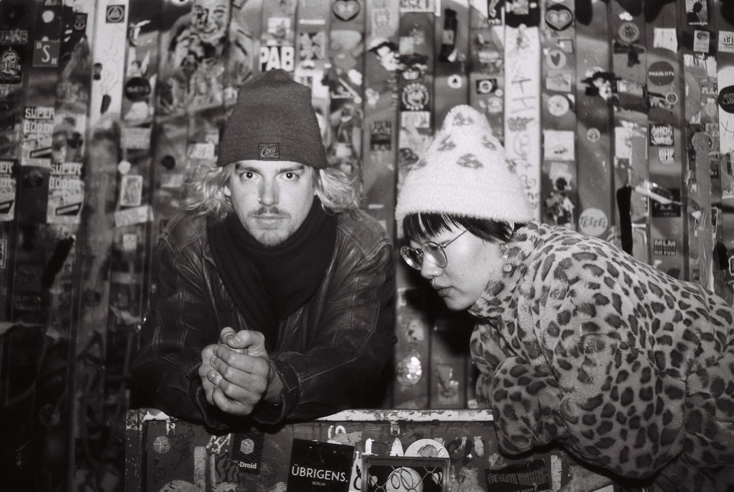 Robin Fisher and Jingyun Li leaning toward the camera in a black-and-white photograph inside a sticker-covered venue.